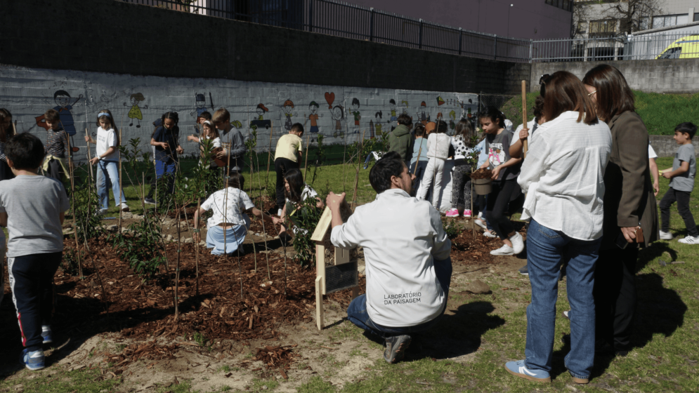 Photo of a community garden activity with children and adults planting and tending to plants in a mulched area, surrounded by grass. A mural with colorful drawings decorates a concrete wall in the background, while participants engage in gardening tasks and observe.