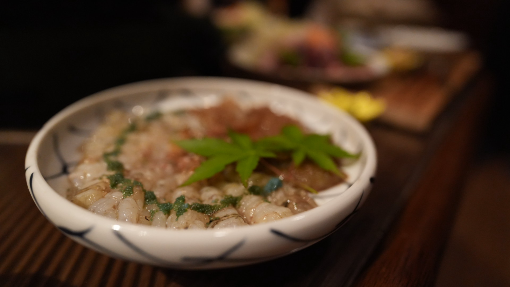 Photo of a white ceramic bowl filled with a dish featuring small, translucent shrimp arranged in a circular pattern and garnished with green leaves. The bowl sits on a wooden surface with a blurred background showing additional food items.