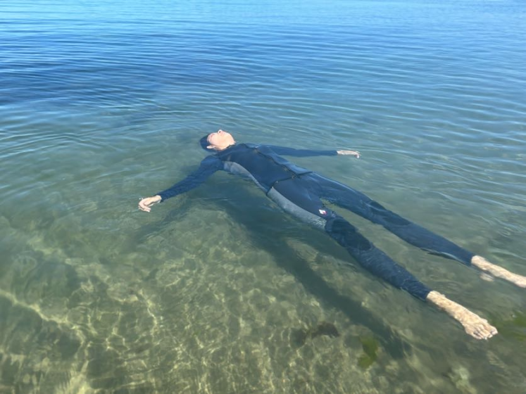 Photo of a person floating on clear, shallow water while wearing a black wetsuit. The calm water reveals a sandy and rocky bottom, with sunlight creating visible shadows and reflections around the floating figure.