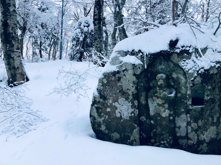 Photo of a snow-covered forest scene featuring a large mossy rock with a carved human figure and a small niche. Surrounding trees and ground are blanketed in fresh snow, creating a serene winter atmosphere.