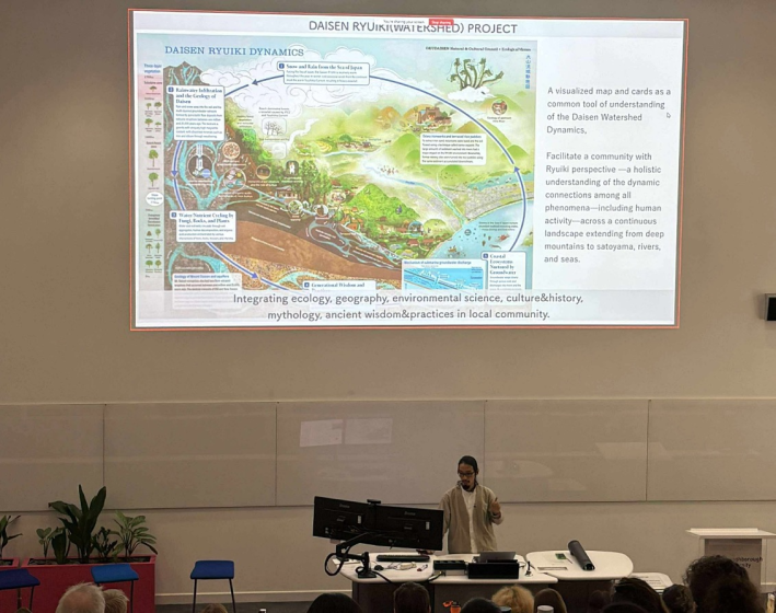 Photo of a lecture presentation showing a detailed watershed project diagram titled "Daisen Ryuki Watershed Project," illustrating ecological, geographical, and cultural dynamics through a colorful map with labeled sections and explanatory text. The diagram highlights integration of environmental science, mythology, and local wisdom, emphasizing connections from mountains to rivers and seas, with key elements like water flow, human activity, and natural features visually represented.