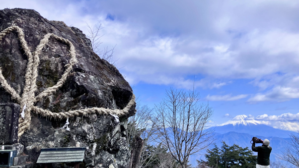 Photo of a person taking a photo of a large rock wrapped with thick, twisted straw ropes, set against a backdrop of leafless trees and snow-capped mountains under a partly cloudy sky. 