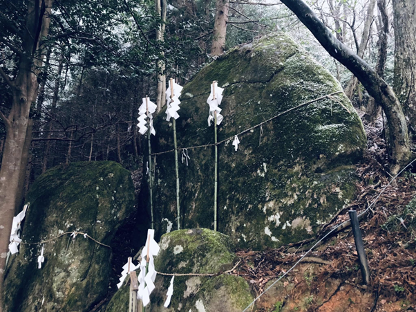 Photo of a natural forest scene featuring large moss-covered rocks adorned with traditional Shinto paper streamers (shide) tied to ropes and bamboo poles. The setup indicates a sacred or ritual site, with white zigzag paper strips symbolizing purification and spiritual presence in Japanese culture.