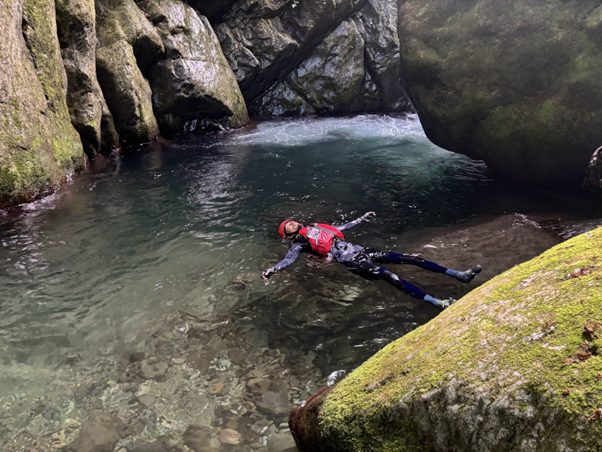 Photo of a person wearing a red life jacket and helmet floating on their back in clear water surrounded by moss-covered rocks and a rocky cave.