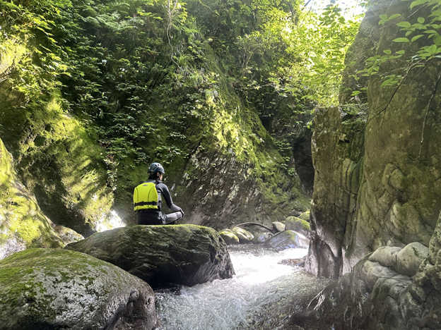 Photo of a person wearing a helmet and yellow life jacket sitting on a moss-covered rock beside a flowing stream in a narrow, lush green canyon