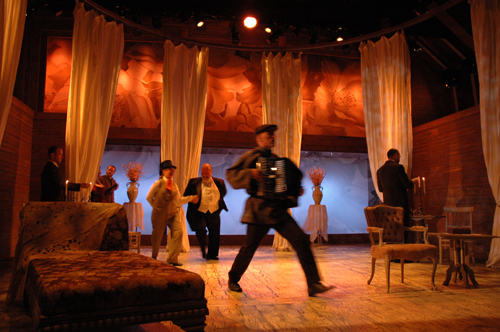 Photograph of a theatrical stage during a dramatic performance featuring actors in period costumes, with one playing an accordion while moving across the stage.