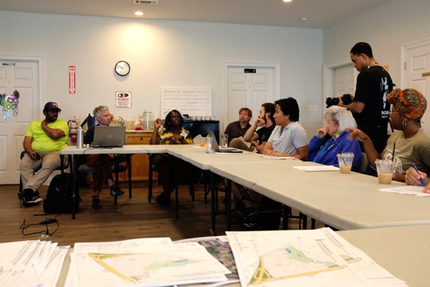 Photo of a community meeting with diverse participants seated around tables arranged in a U-shape, discussing documents and maps spread out in the foreground. The room features neutral walls, a clock, and a motivational poster