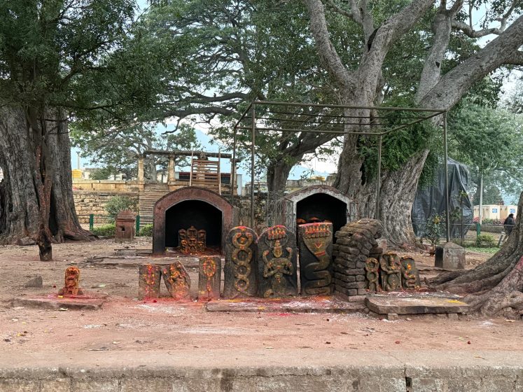 Photograph of an outdoor kiln setup with multiple brick and clay sculptures arranged in front, some covered in red pigment. Large trees surround the area, and two brick kilns with arched openings are visible in the background, indicating a traditional pottery or ceramic firing site.