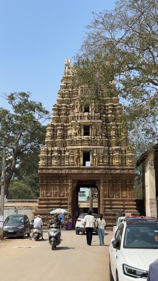Photo of an ancient temple entrance tower with intricate stone carvings, surrounded by trees and vehicles. The scene captures daily activity with people walking and riding scooters near the historic structure under a clear blue sky.