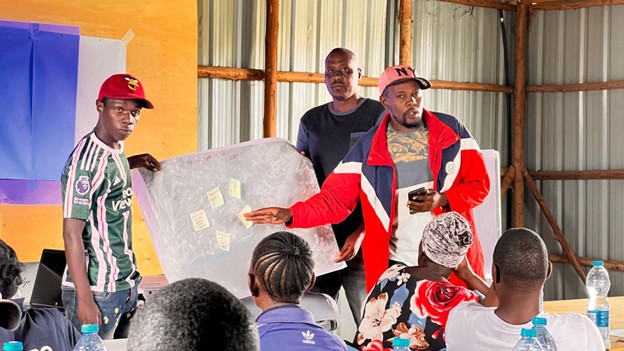 Photo of a group meeting inside a metal-walled room where one person in a red jacket is presenting a transparent board with sticky notes to seated attendees