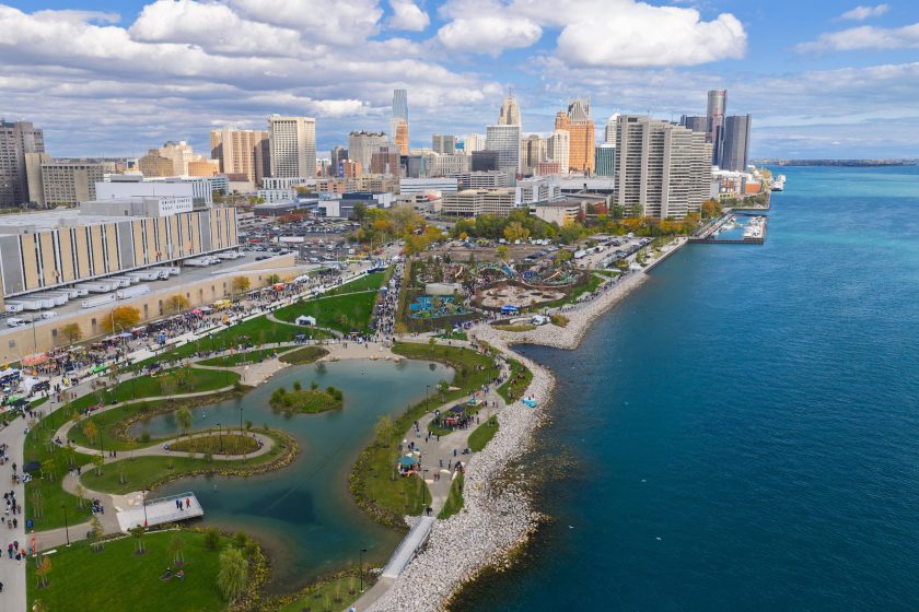 Aerial photograph of an urban waterfront park with winding walking paths, small ponds, and green spaces adjacent to a large body of water. City skyline with tall buildings and partly cloudy sky forms background