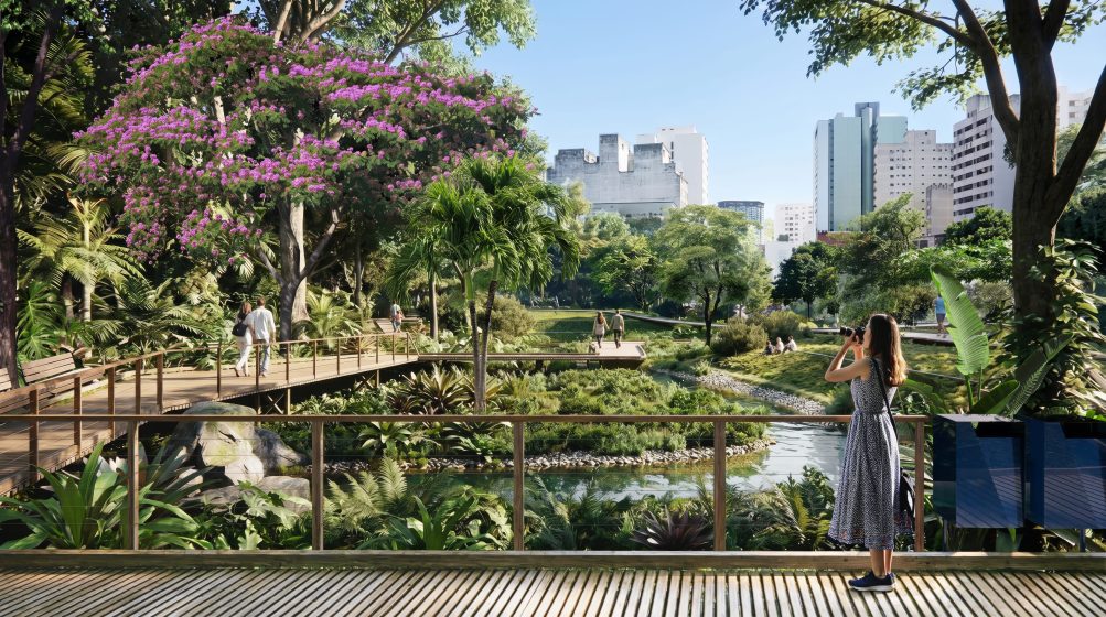 Photo of an urban park featuring a wooden boardwalk, lush greenery, and a small waterway with people walking and relaxing. Tall buildings in background contrast with vibrant trees, including one with bright pink flowers