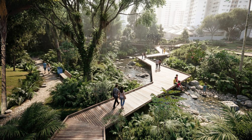 Photo of a lush urban park featuring a wooden boardwalk crossing over a small stream with rocks and greenery. Several people are walking and sitting along the path, surrounded by dense trees and plants, with tall buildings visible in the background.