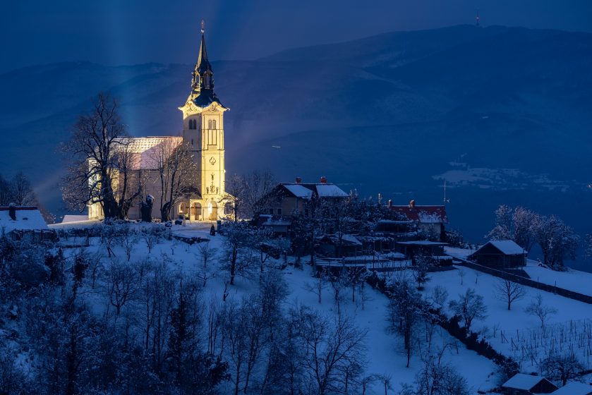 Photo of a snow-covered village at night featuring a brightly illuminated church with a tall steeple as the focal point. Surrounding buildings and leafless trees are dimly visible against a backdrop of dark blue mountains and sky, creating a serene winter scene.