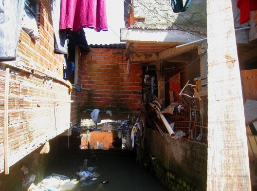 Photo of a narrow, cluttered alleyway between brick buildings with stagnant water filled with debris and trash. Clothes hang overhead, and various discarded items, including pipes and metal objects, are scattered along walls and water