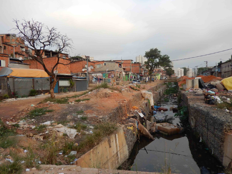 Photo of an urban slum area showing makeshift housing made of brick and metal sheets alongside a polluted, narrow canal filled with debris and stagnant water. The scene includes a leafless tree, scattered trash, and distant high-rise buildings under a cloudy sky