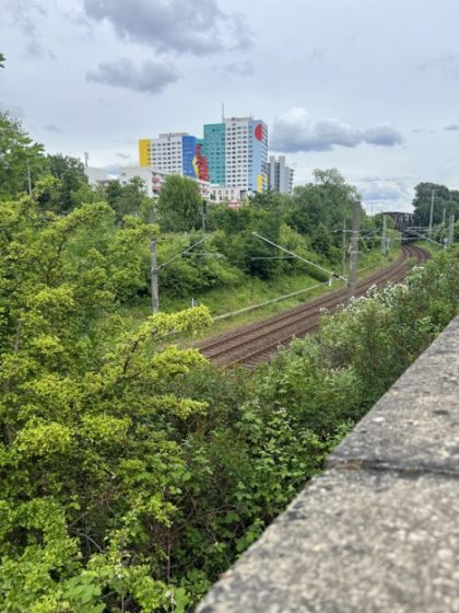 Photo showing a curved railway track surrounded by dense green foliage and trees under a cloudy sky. In the background, colorful high-rise buildings with murals stand out, adding an urban contrast to the natural and industrial elements in the scene.