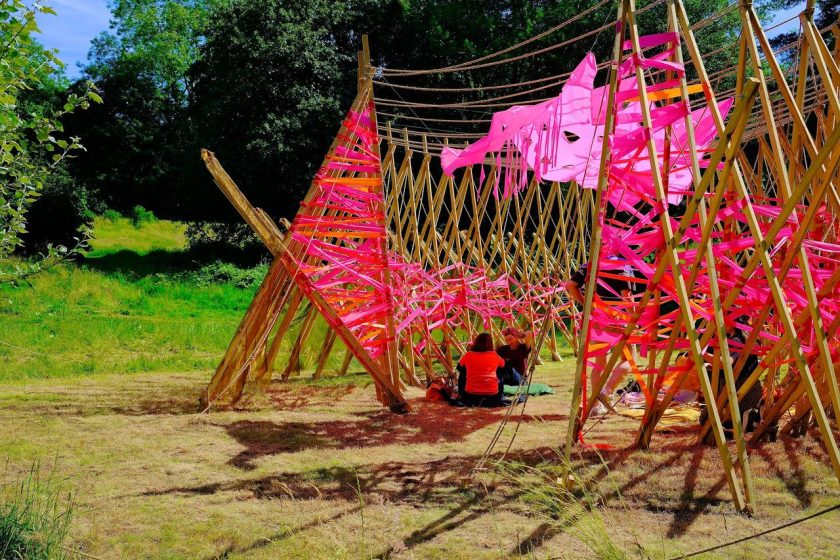 Photo of an outdoor art installation featuring a wooden frame structure wrapped with bright pink fabric strips, creating a tunnel-like effect. Two people sit inside the structure on grass, surrounded by green trees and sunlight casting shadows on the ground.