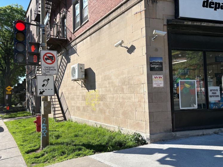 Photo of a street corner featuring a red traffic light, pedestrian signal with a countdown timer, and multiple street signs including a no left turn sign and an accessibility sign with a hearing aid symbol. The corner shows a brick building with security cameras, an air conditioning unit, and a storefront window displaying posters.
