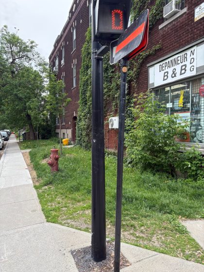 Photo of a street corner featuring a pedestrian crossing signal displaying a red hand with a countdown of zero. Surrounding elements include a brick building with a "DEPANNEUR B & G" sign, a fire hydrant, sidewalk, and greenery along the street.