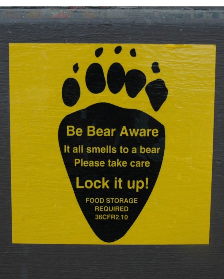 Warning sign with a black bear paw print on yellow background advising to secure food storage to prevent attracting bears. Text emphasizes caution with phrases "Be Bear Aware," "Lock it up!," and references food storage regulation 36CFR2.10.