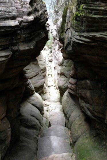 Photo of a narrow rocky canyon with steep, layered walls showing natural erosion patterns. Light filters through the top, 