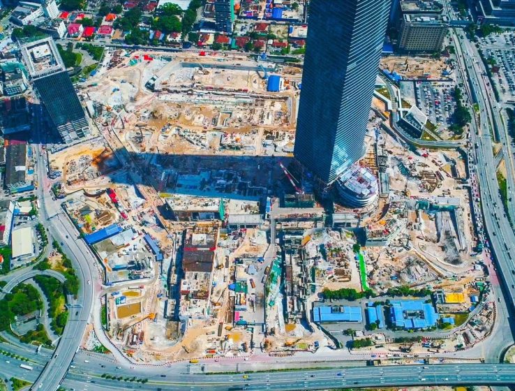Aerial photograph of a large urban construction site featuring multiple building foundations and cranes surrounding a tall, completed skyscraper. The site is bordered by busy roads and includes areas with construction materials, vehicles, and temporary blue-roofed structures.