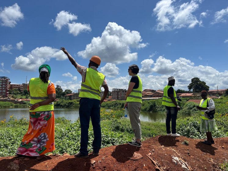 Photograph of five people wearing yellow safety vests standing on a dirt mound near a body of water with greenery and buildings in the background. One person gestures toward the sky under a partly cloudy blue sky