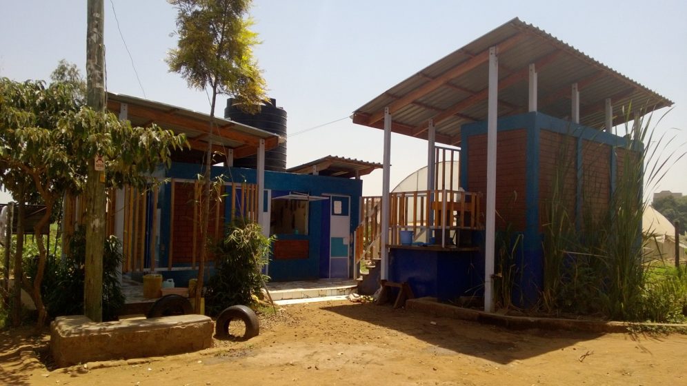 Photo of a small outdoor facility with two blue and brown structures featuring wooden railings and metal roofs, set in a dry, dusty environment with some vegetation. The scene includes a water tank on one building, a tree, and various plants
