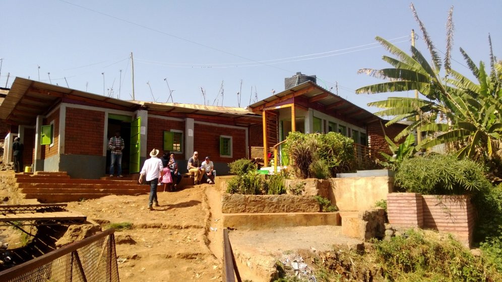 Photo of a rural house with red brick walls and green window frames, situated on a dirt path with several people standing and sitting outside. Surrounding area includes plants, banana trees, and a clear blue sky, indicating a warm, sunny day in a countryside setting.