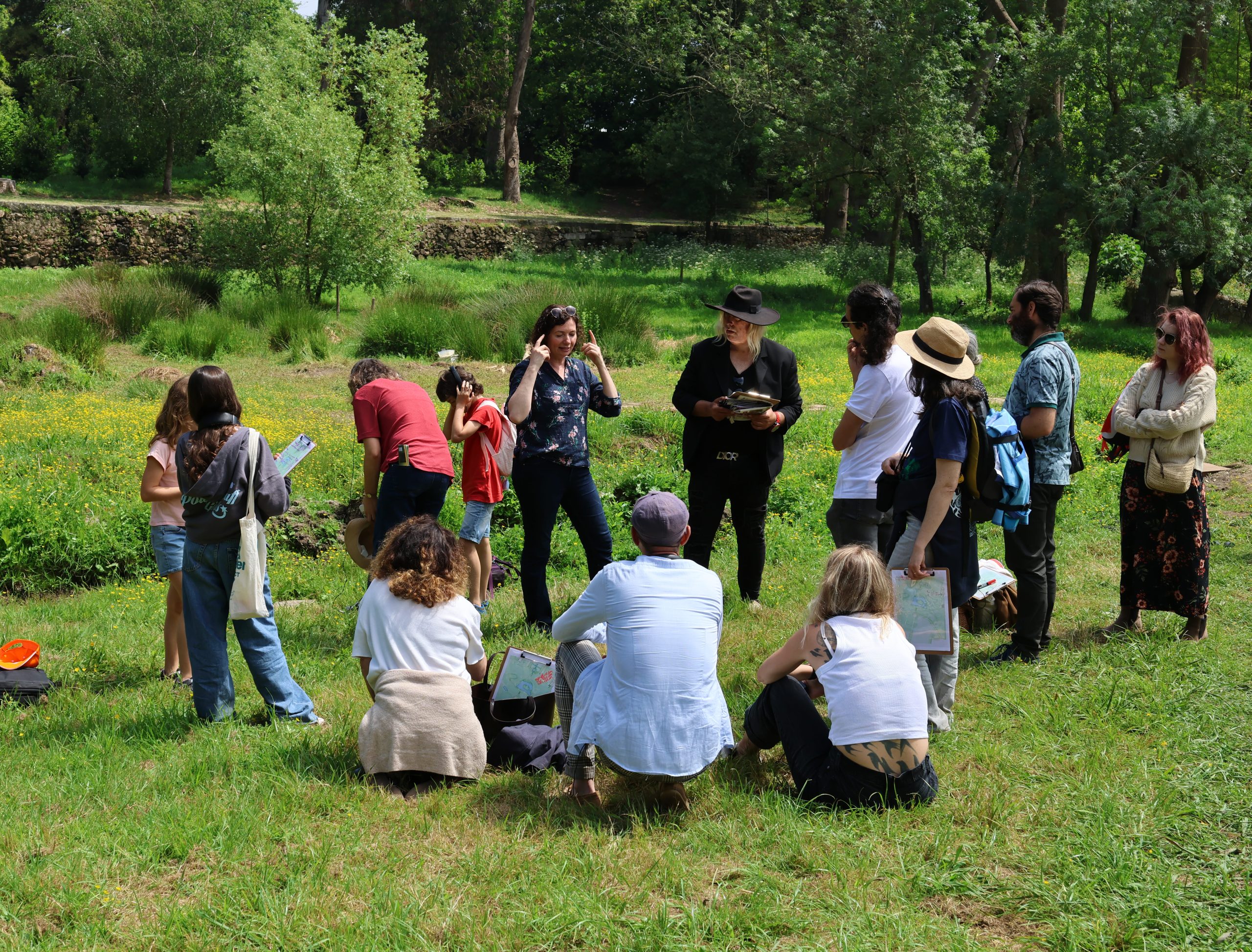 Photo of a group of people gathered outdoors in a grassy area, likely participating in an educational or nature-related activity. The group includes individuals standing and sitting, with some holding notebooks or papers, surrounded by trees and greenery under natural daylight.