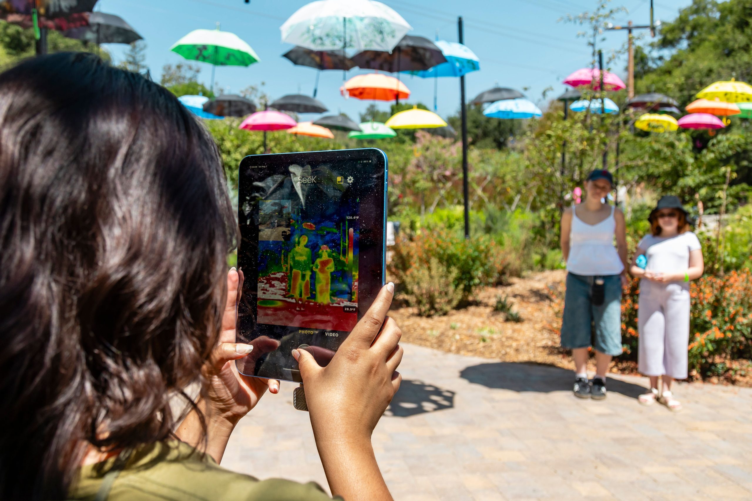 Photo showing a person using a thermal imaging device to capture two individuals standing outdoors under colorful umbrellas.