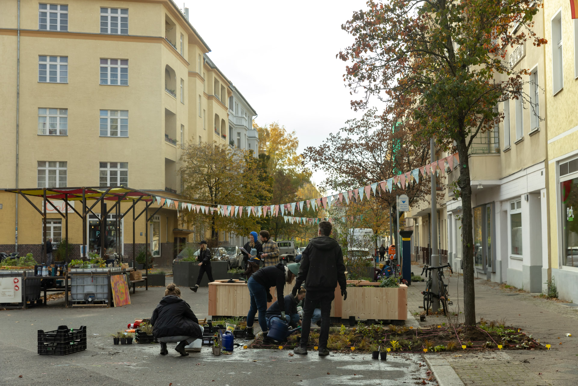 Photo of a street scene showing several people planting flowers and arranging plants in an urban area between apartment buildings. Colorful triangular flags hang overhead