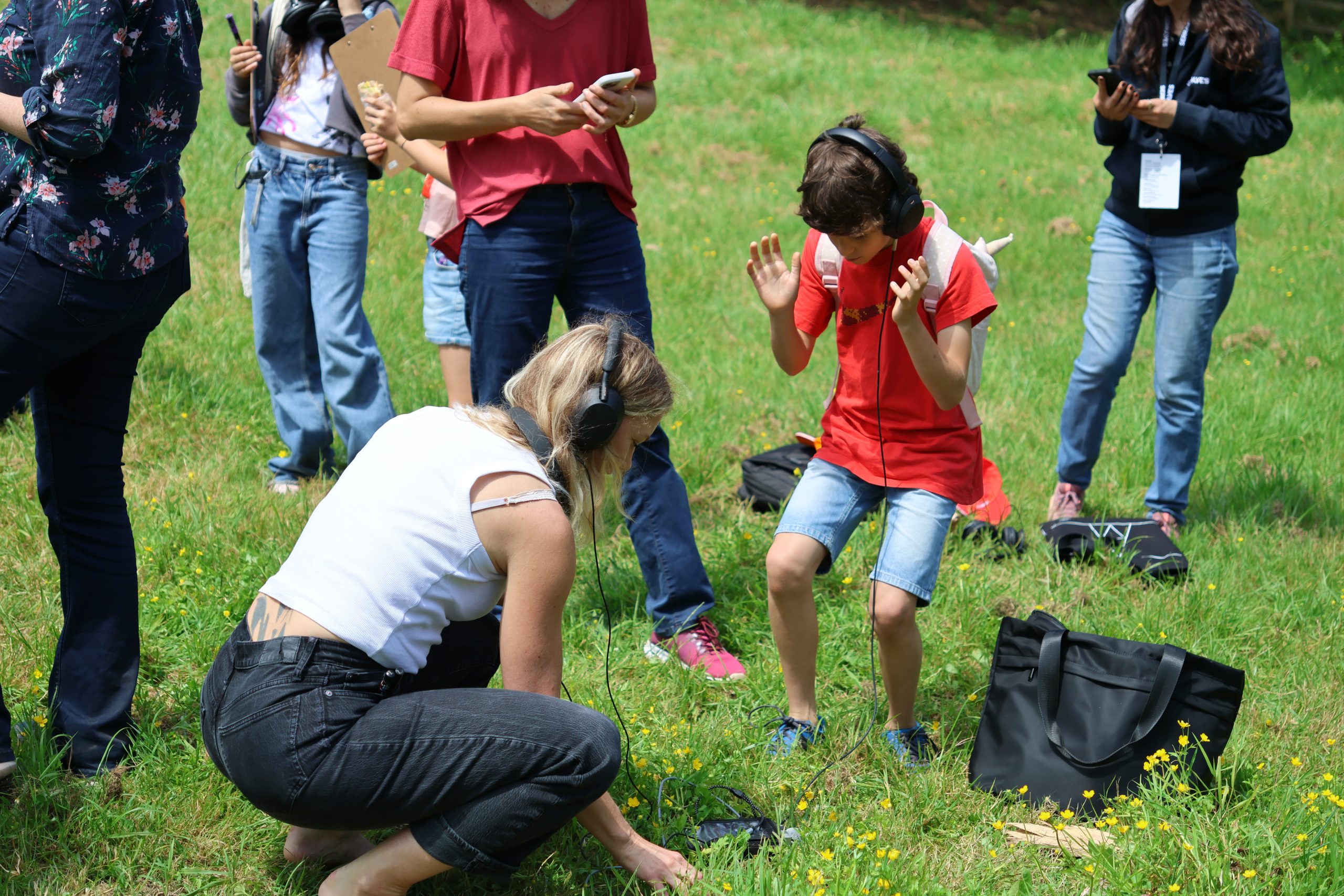 Photoshowing a group of people outdoors on grass, with a woman crouching and a child standing, both wearing headphones and seemingly engaged in an audio activity. Other individuals stand nearby, some holding devices, suggesting a group event or workshop focused on listening or sound exploration.