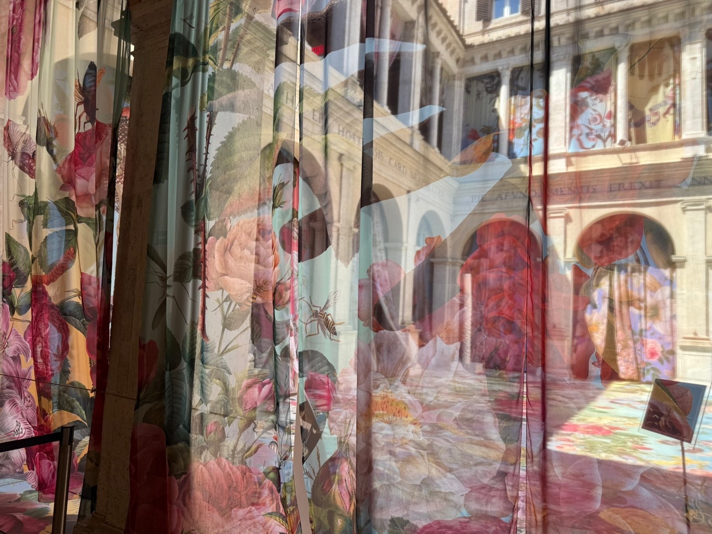 Photo of a mural showing a floral-patterned curtain with pink and red roses partially transparent against a background of a historic building with arches and columns. 