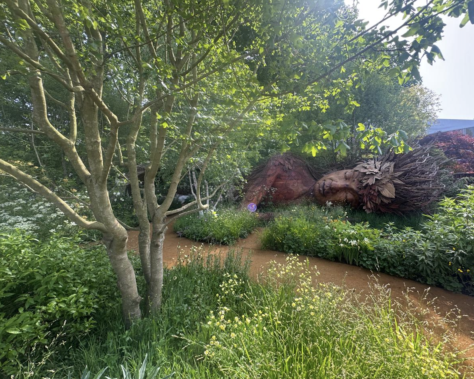 Photo of a tree in a grassy field on the edge of a darkened forest. In the forest, a giant wooden statue of a figure lays sleeping amongst the trees.