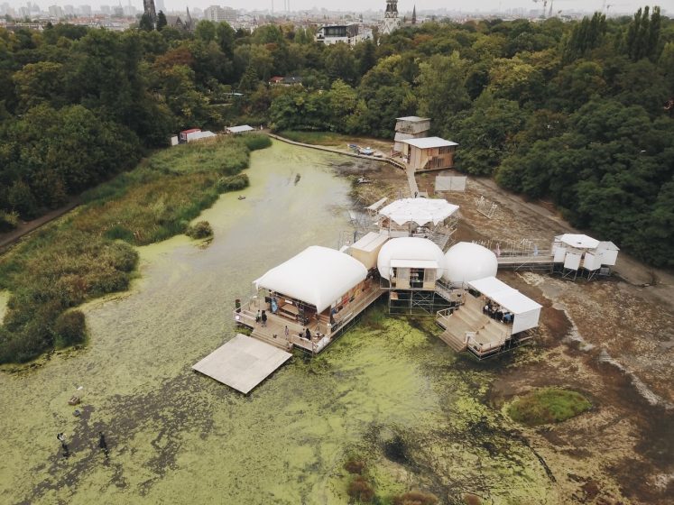 Aerial photo of a floating structure complex on a partially algae-covered body of water surrounded by dense trees. The complex includes multiple connected platforms with white dome and tent-like coverings, wooden walkways, and small buildings