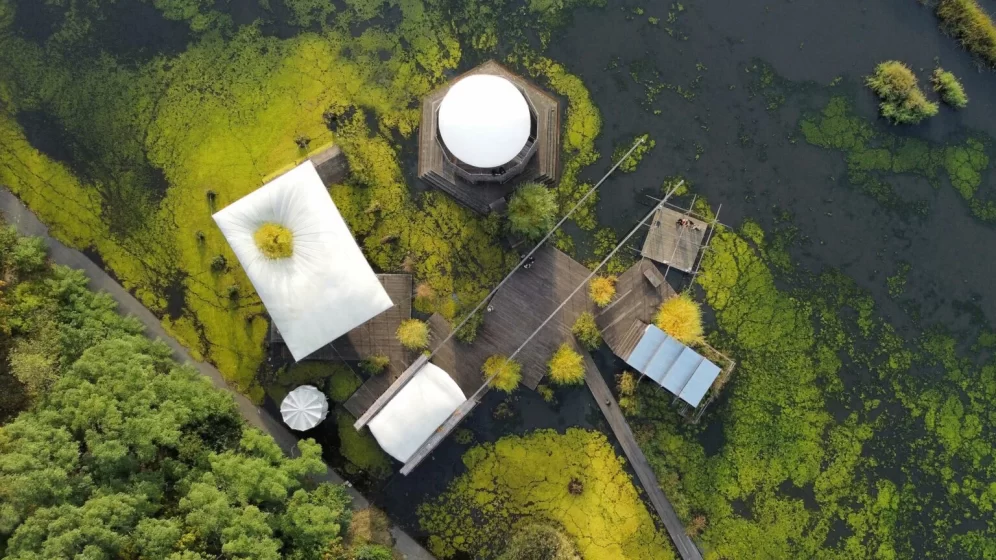 Aerial photo showing a cluster of buildings and wooden walkways surrounded by green algae-covered water and dense vegetation. The structures include a large circular white dome, rectangular white roofs, and a small dock area, highlighting a remote or aquatic research or recreational facility.
