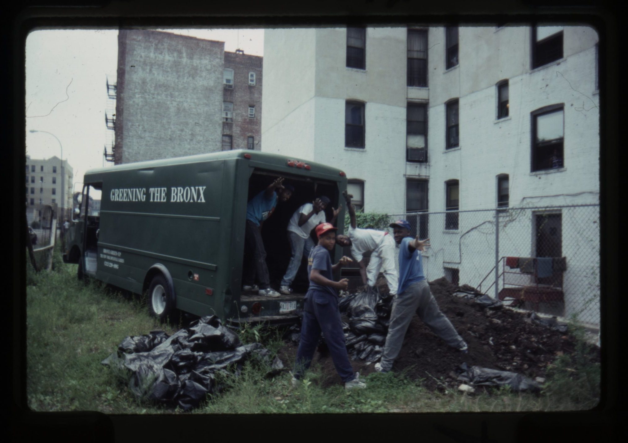 Photograph showing workers loading black garbage bags into a green van labeled "New York City Sanitation." The scene takes place in an urban area with a white multi-story building and a chain-link fence in the background.
