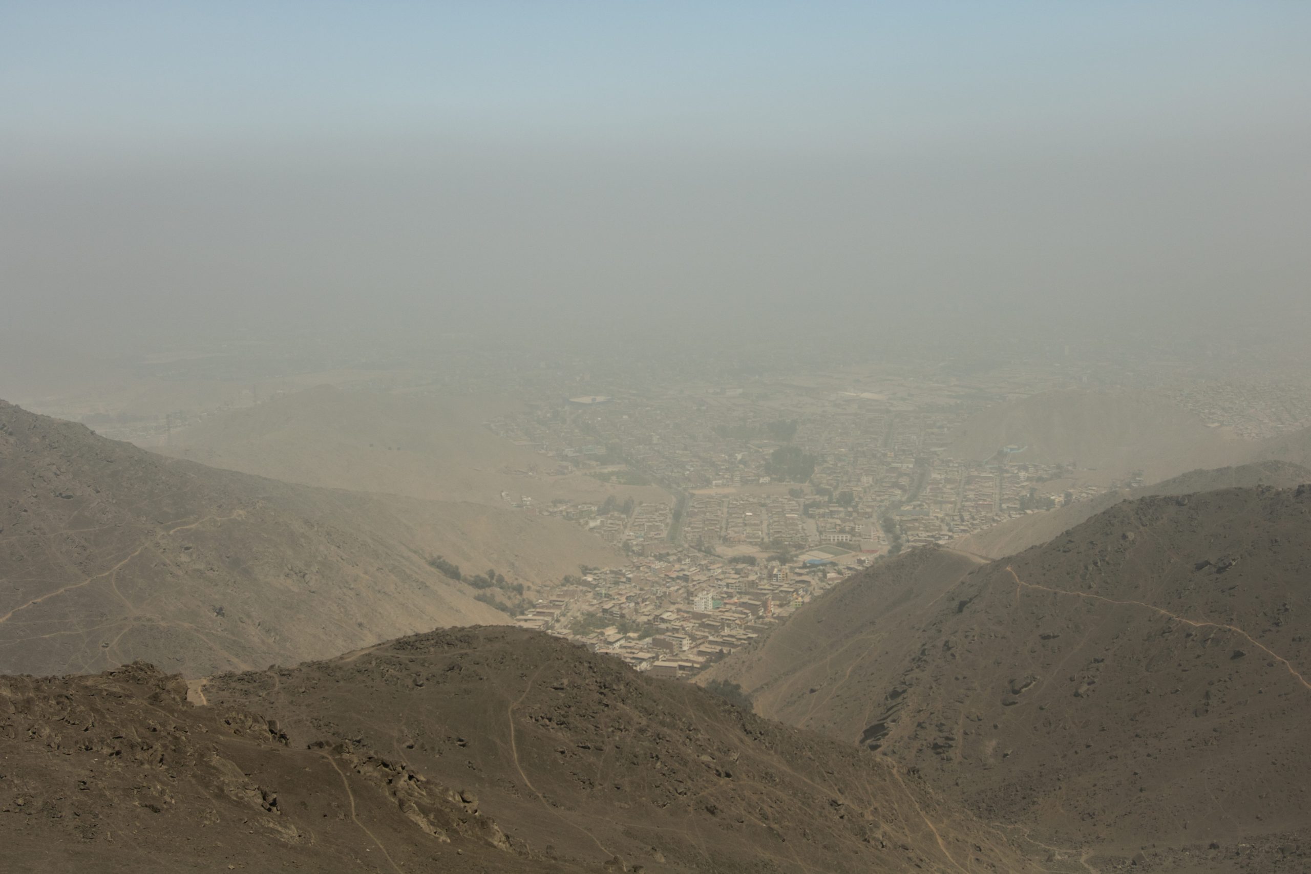 Photograph showing a hazy view of a city nestled between brown, barren hills. Visibility is low due to dust or pollution, obscuring detailed features of urban structures and landscape.