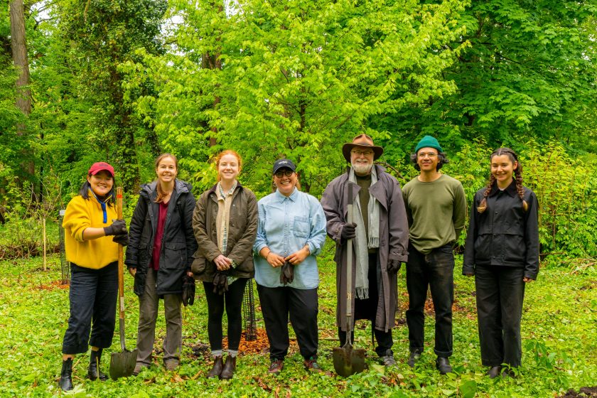 Photo of seven people standing outdoors in a green, wooded area, each holding a shovel or standing with hands by their sides. Group wears casual outdoor clothing, including jackets, hats, and boots
