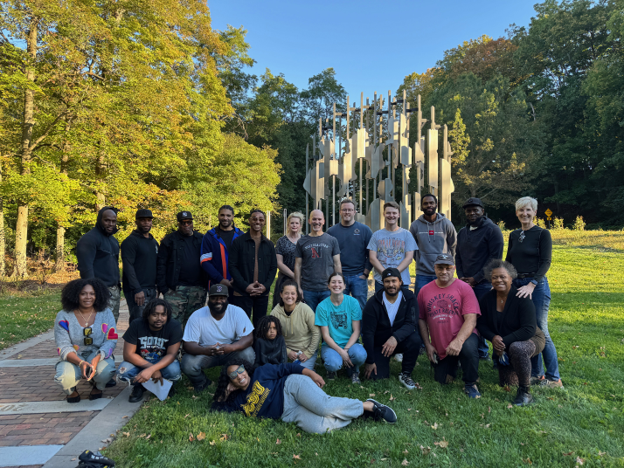 Photo of a diverse group of 21 people posing outdoors on grass in front of a tall, abstract metal sculpture surrounded by trees with autumn foliage. Group members display casual attire, smiling expressions, and a relaxed atmosphere