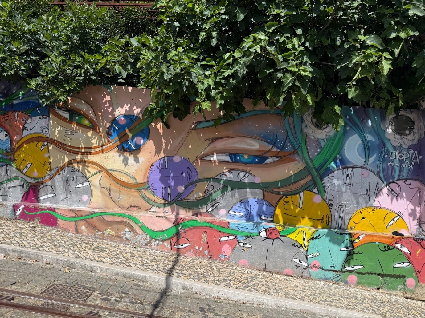 Colorful street mural depicting a stylized human face with closed eyes surrounded by abstract shapes and vibrant orbs in yellow, purple, blue, and green. The mural is painted on a concrete wall partially covered by leafy tree branches, with a cobblestone sidewalk and tram tracks in front.