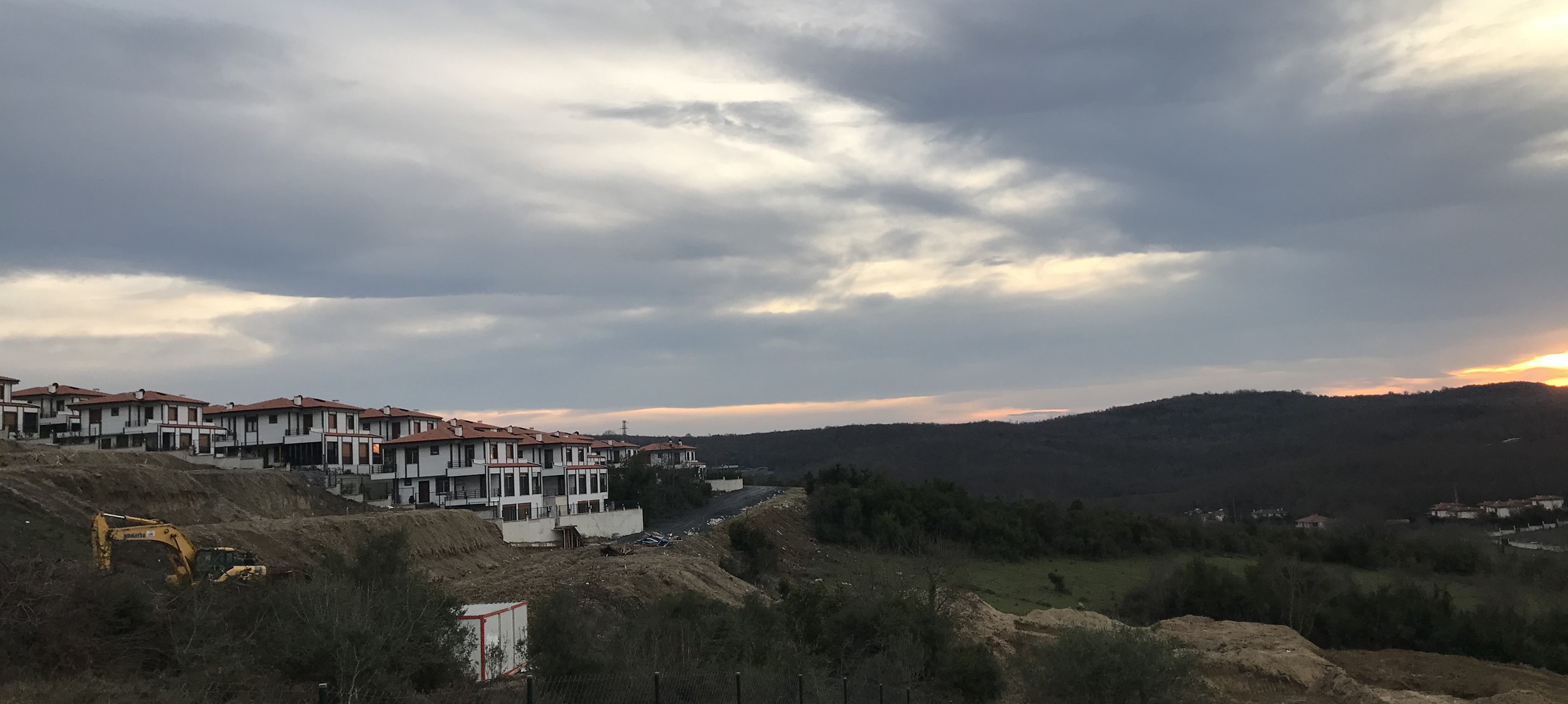 Photo of a residential construction site on a hillside during sunset, showing partially built white houses with red roofs on the left and green forested hills extending to the right. The sky is mostly cloudy with a bright area near the horizon