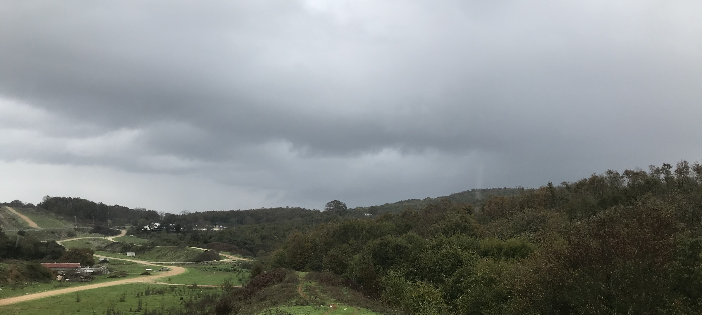 Photo of a rural landscape featuring rolling hills, dense green trees, and a winding dirt road leading to scattered houses. Overcast sky with thick gray clouds suggests an approaching storm or rainy weather.