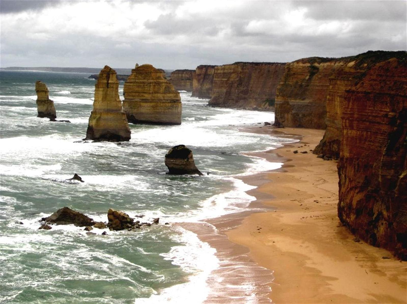Photo of a coastal landscape featuring tall limestone stacks rising from ocean waves along a sandy beach bordered by rugged cliffs. The scene highlights natural erosion processes with cloudy skies overhead and scattered rock formations in the water.