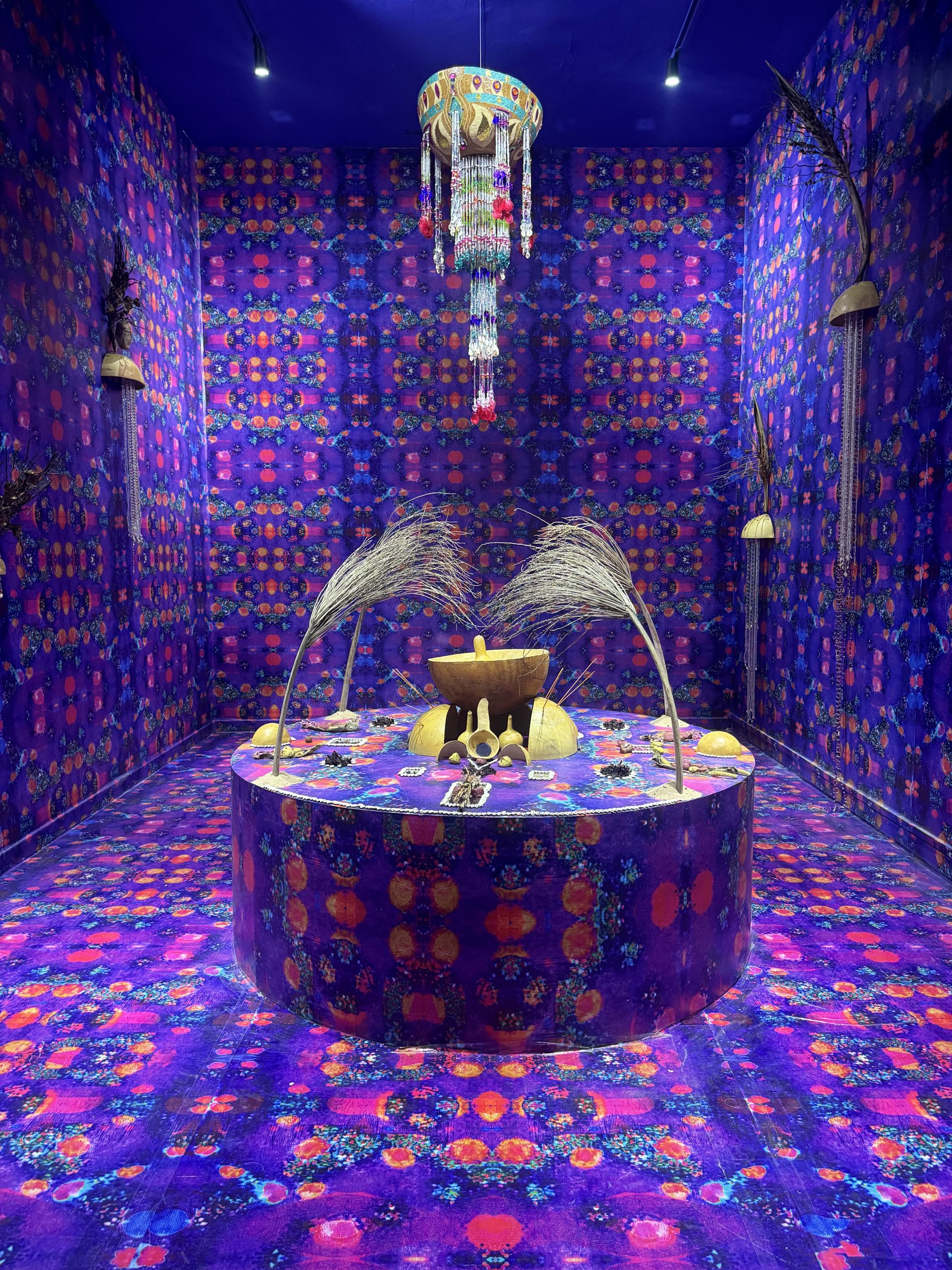 Photo of an art installation featuring a circular platform covered in vibrant purple and orange patterned fabric, surrounded by matching walls and floor. The platform displays various objects including dried grasses, pottery, and small sculptures, with a colorful hanging mobile suspended above, creating a cohesive and immersive visual environment.