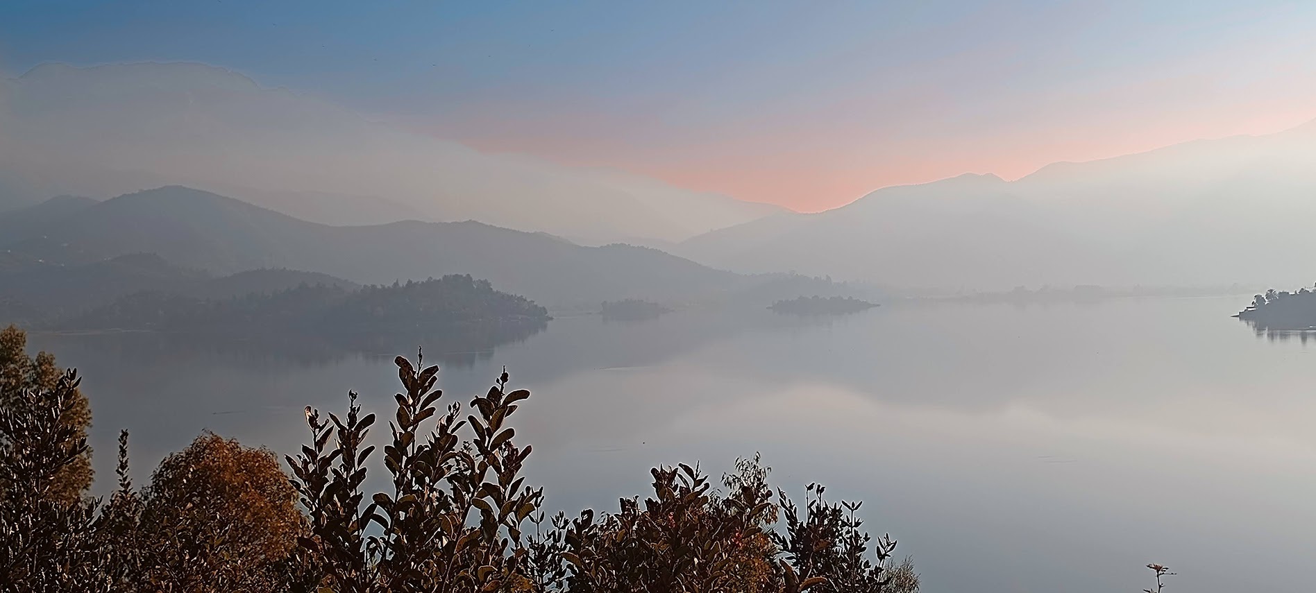 Photo of a mist-covered mountain lake surrounded by layered hills and silhouetted foreground foliage. Soft sunrise colors, calm reflective water, and distant islands create a tranquil landscape scene.