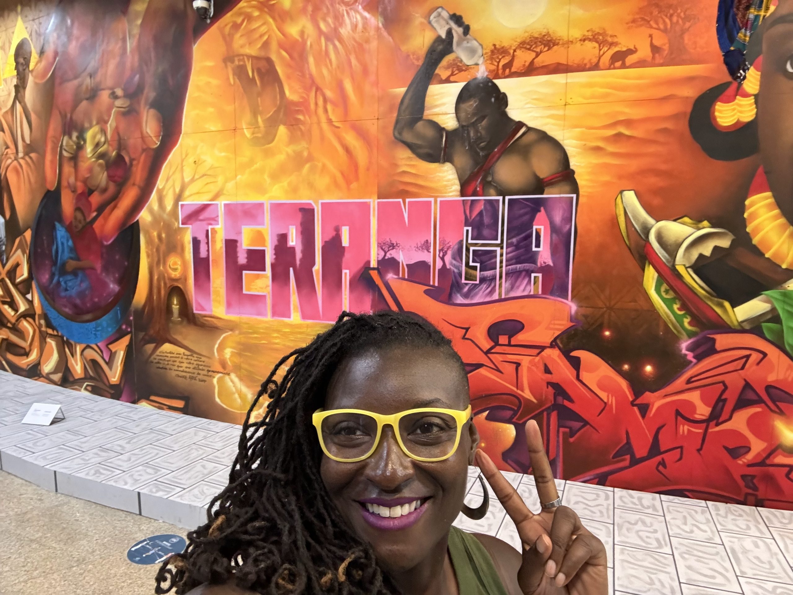 Photo of a smiling person wearing bright yellow glasses posing with a peace sign in front of a colorful mural. Mural features bold text "TERANGA," vibrant red and orange hues, and artistic depictions of cultural and historical themes.