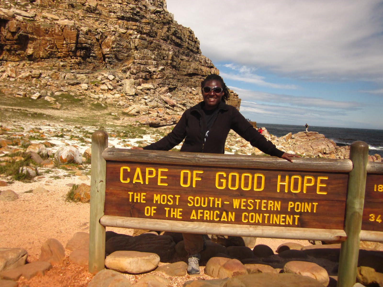 Photo of a person standing behind a wooden sign at Cape of Good Hope, indicating it as the most south-western point of the African continent. Rocky terrain and a partly cloudy sky form the background